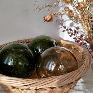 Peut inclure: Un panier en osier tressé contient trois sphères de verre vertes et ambrées. Le panier est posé sur une nappe blanche avec des broderies florales. Des herbes et des fleurs séchées sont en arrière-plan.