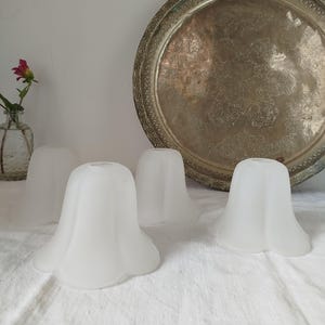 May include: Four frosted glass lampshades with a tulip-like shape are arranged on a white cloth. A large, ornate metal tray sits in the background. A small glass vase with a pink flower is on the left.