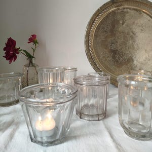 以下が含まれることがあります： 透明なガラス製のキャンドルホルダーのコレクション。そのうちの1つにはろうそくが灯っています。ヴィンテージのシルバートレイと赤い花が入った小さなガラスの花瓶が背景にあります。キャンドルホルダーは白い布の上にあります。