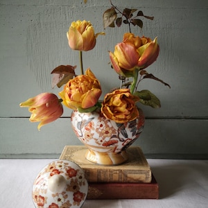 May include: A ceramic vase with a floral pattern and a lid, filled with orange and yellow tulips. The vase is sitting on top of two books.
