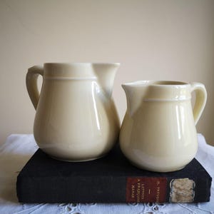 May include: Two cream-colored ceramic pitchers with handles, one larger than the other, sit on a dark book. The pitchers have a classic design and are placed on a white lace tablecloth.