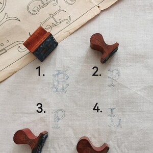 May include: A collection of vintage wooden stamps with red handles and black inked surfaces. The stamps have printed the letters B, P, and U in light blue ink on a white fabric. A vintage paper with ornate letter designs is in the background.