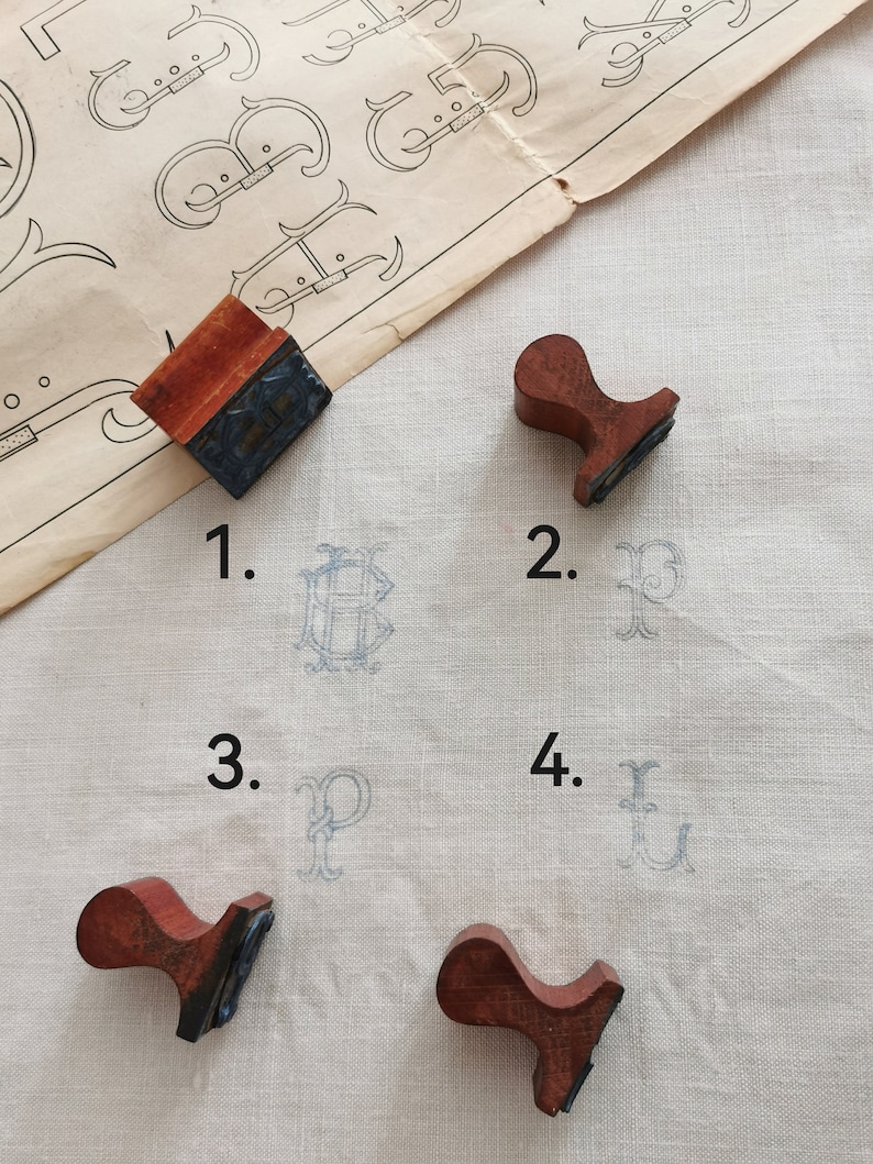 May include: A collection of vintage wooden stamps with red handles and black inked surfaces. The stamps have printed the letters B, P, and U in light blue ink on a white fabric. A vintage paper with ornate letter designs is in the background.