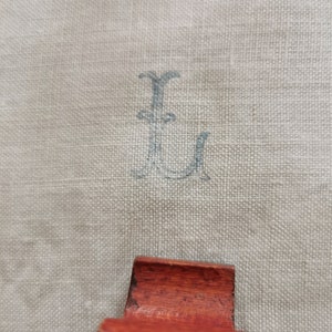May include: Close-up of a light beige linen fabric with a faded blue monogram letter "L" printed on it. A small, reddish-brown wooden object is in the foreground.