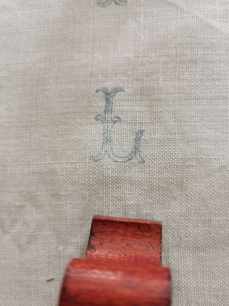 May include: Close-up of a light beige linen fabric with a faded blue monogram letter "L" printed on it. A small, reddish-brown wooden object is in the foreground.