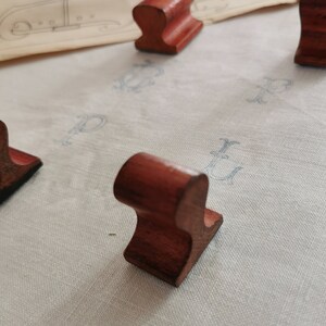 May include: Several small, reddish-brown wooden objects with a curved top and a flat base are arranged on a light-colored surface. The objects appear to be handmade and have a smooth finish. The background includes a vintage paper with faded blue markings.