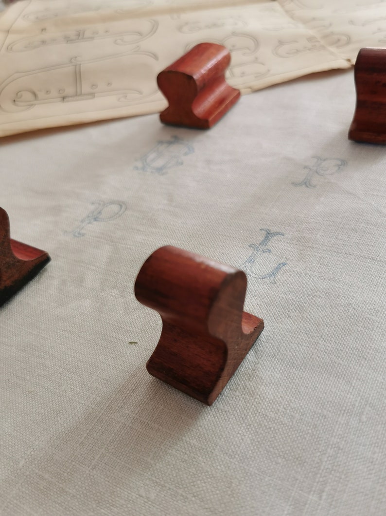 May include: Several small, reddish-brown wooden objects with a curved top and a flat base are arranged on a light-colored surface. The objects appear to be handmade and have a smooth finish. The background includes a vintage paper with faded blue markings.