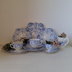 Op de afbeelding: Een vintage theeservies met een blauw en wit bloemenpatroon. De set bevat een theepot, roomkan, suikerpot, twee theekopjes en schoteltjes. De theekopjes en schoteltjes hebben gegolfde randen.