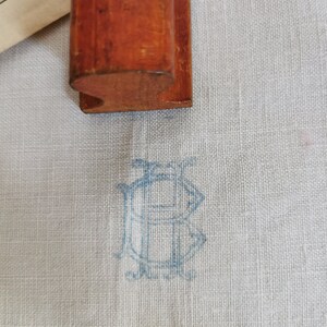 May include: A wooden stamp with a curved base rests on a light blue fabric. The fabric features a faded blue monogram design. The stamp is a warm brown color, and the fabric has a subtle texture.