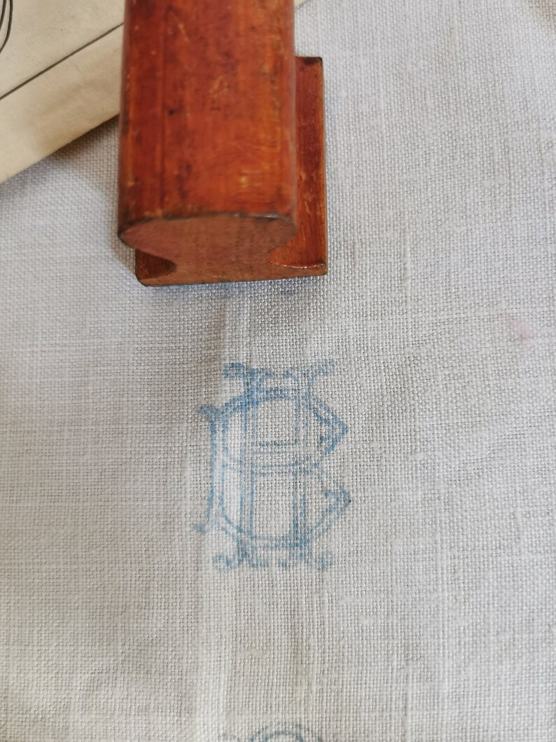 May include: A wooden stamp with a curved base rests on a light blue fabric. The fabric features a faded blue monogram design. The stamp is a warm brown color, and the fabric has a subtle texture.