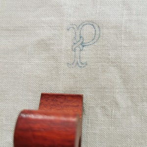 May include: A dark reddish-brown wooden napkin ring with a curved front and a rectangular back. The background is a light gray linen fabric with a blue embroidered letter "P".