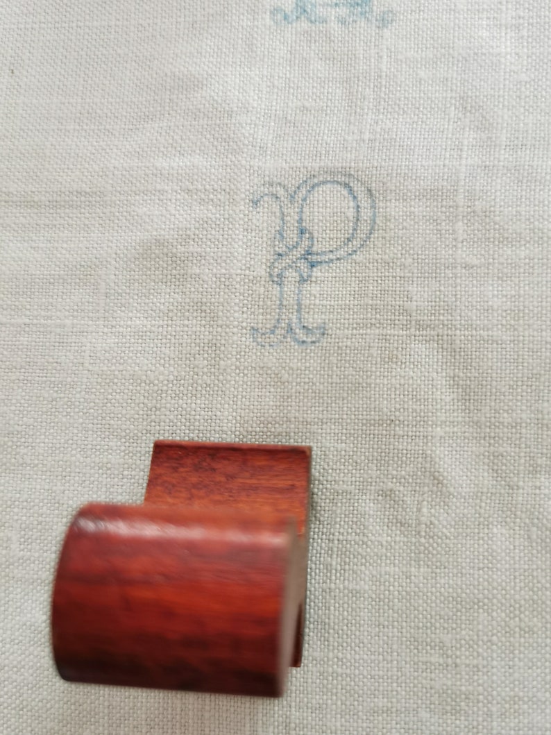 May include: A dark reddish-brown wooden napkin ring with a curved front and a rectangular back. The background is a light gray linen fabric with a blue embroidered letter "P".