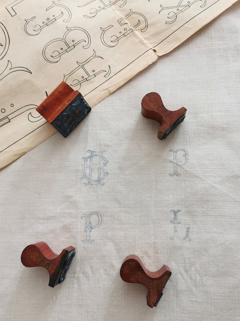 May include: An overhead shot shows a collection of vintage letter stamps with wooden handles and inked surfaces. The stamps are arranged on a white linen surface with faint blue letter imprints. A sheet of paper with letter designs is in the background.