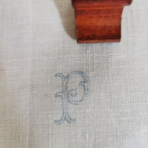 May include: Close-up of a light-colored linen fabric with a delicate, embroidered blue letter "P". A dark wood element is partially visible at the top. The fabric has a subtle texture, and the embroidery adds a touch of elegance. The image suggests a focus on textiles and personalization.