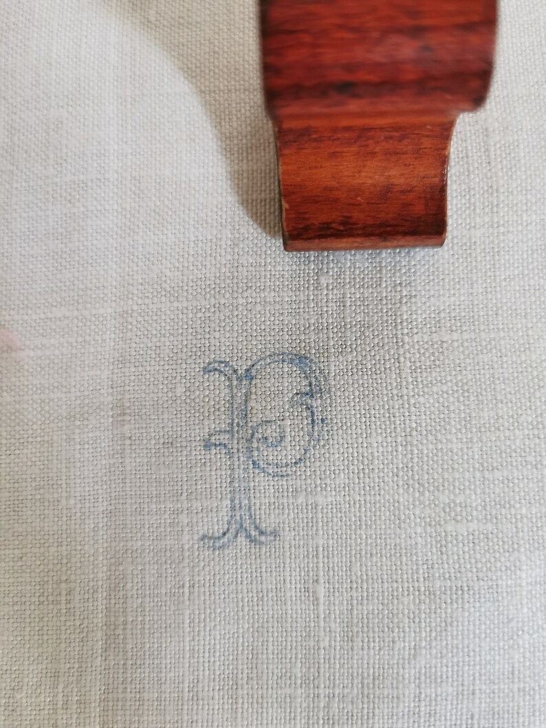 May include: Close-up of a light-colored linen fabric with a delicate, embroidered blue letter "P". A dark wood element is partially visible at the top. The fabric has a subtle texture, and the embroidery adds a touch of elegance. The image suggests a focus on textiles and personalization.