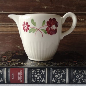 Op de afbeelding: Een witte keramische roomkan met een bloemenmotief in rood en groene tinten. De roomkan heeft een geribbeld lichaam en een handvat. Hij staat op een zwart boek met gouden letters waarop "Contes Licencieux" en "Sade" staat.