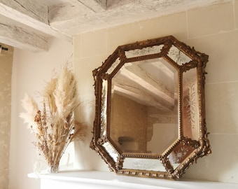 May include: A large, ornate, gold-framed mirror with a beveled, octagonal glass. The mirror is hanging on a white wall above a white mantelpiece. The mirror reflects the white ceiling beams and a bouquet of dried pampas grass.