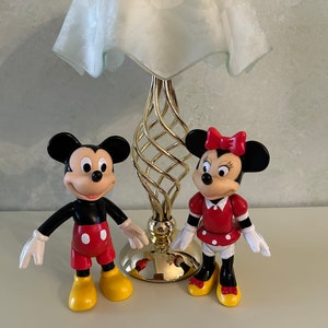 May include: A gold table lamp with a white frosted glass shade and a pair of Mickey Mouse and Minnie Mouse figurines. Mickey Mouse is wearing red shorts and yellow shoes. Minnie Mouse is wearing a red dress with white polka dots and yellow shoes.