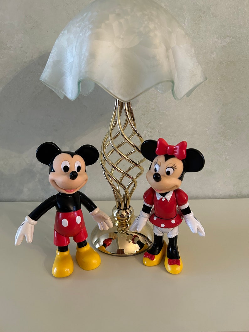 May include: A gold table lamp with a white frosted glass shade and a pair of Mickey Mouse and Minnie Mouse figurines. Mickey Mouse is wearing red shorts and yellow shoes. Minnie Mouse is wearing a red dress with white polka dots and yellow shoes.