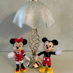 May include: A gold table lamp with a white frosted glass shade and a spiral base.  Two plastic figurines of Mickey Mouse and Minnie Mouse are in front of the lamp. Mickey Mouse is wearing red shorts and yellow shoes. Minnie Mouse is wearing a red dress with white polka dots and red shoes.