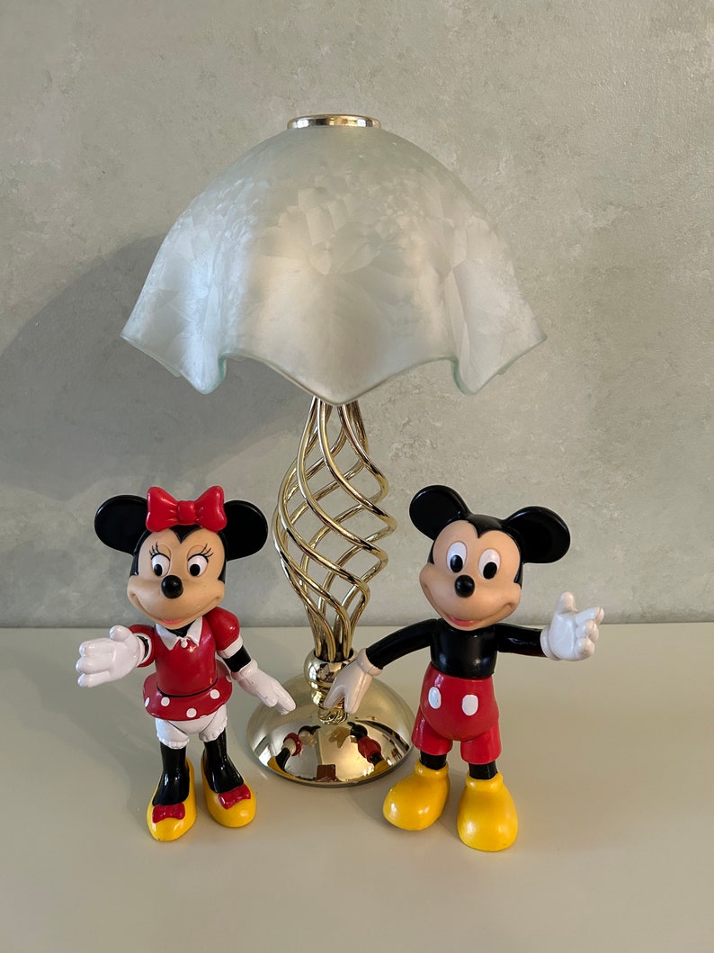 May include: A gold table lamp with a white frosted glass shade and a spiral base.  Two plastic figurines of Mickey Mouse and Minnie Mouse are in front of the lamp. Mickey Mouse is wearing red shorts and yellow shoes. Minnie Mouse is wearing a red dress with white polka dots and red shoes.