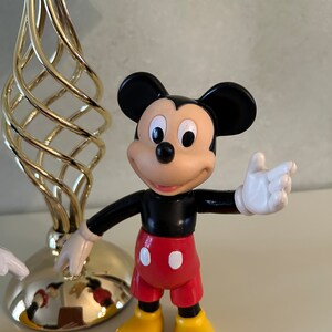May include: A black and red plastic Mickey Mouse figurine with yellow shoes and a white glove. The figurine is standing on a white surface.