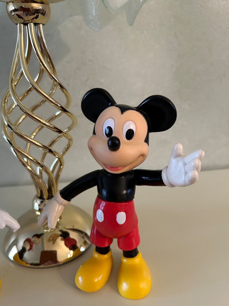 May include: A black and red plastic Mickey Mouse figurine with yellow shoes and a white glove. The figurine is standing on a white surface.