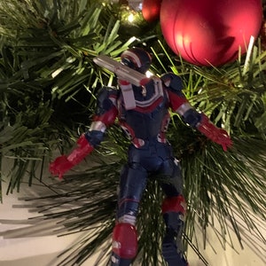 May include: A red, white and blue action figure of a superhero wearing a helmet and holding a weapon. The figure is hanging from a Christmas tree with red and gold baubles.