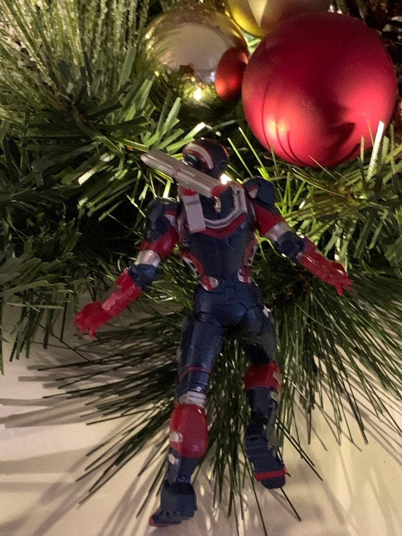 May include: A red, white and blue action figure of a superhero wearing a helmet and holding a weapon. The figure is hanging from a Christmas tree with red and gold baubles.