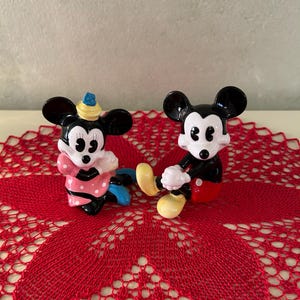 May include: Two ceramic figurines of Mickey and Minnie Mouse. Mickey is in a seated position with red shorts, yellow shoes, and white gloves. Minnie is wearing a pink polka-dot dress and a hat. They are on a red lace doily.