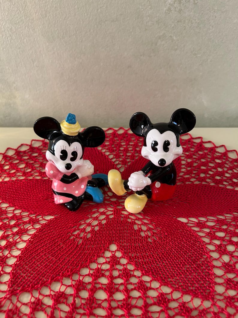 May include: Two ceramic figurines of Mickey and Minnie Mouse. Mickey is in a seated position with red shorts, yellow shoes, and white gloves. Minnie is wearing a pink polka-dot dress and a hat. They are on a red lace doily.
