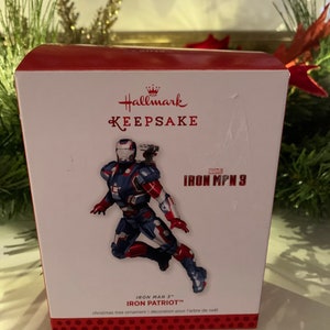 May include: Red and white Hallmark Keepsake ornament box featuring Iron Patriot from Iron Man 3. The ornament is a Christmas tree decoration.