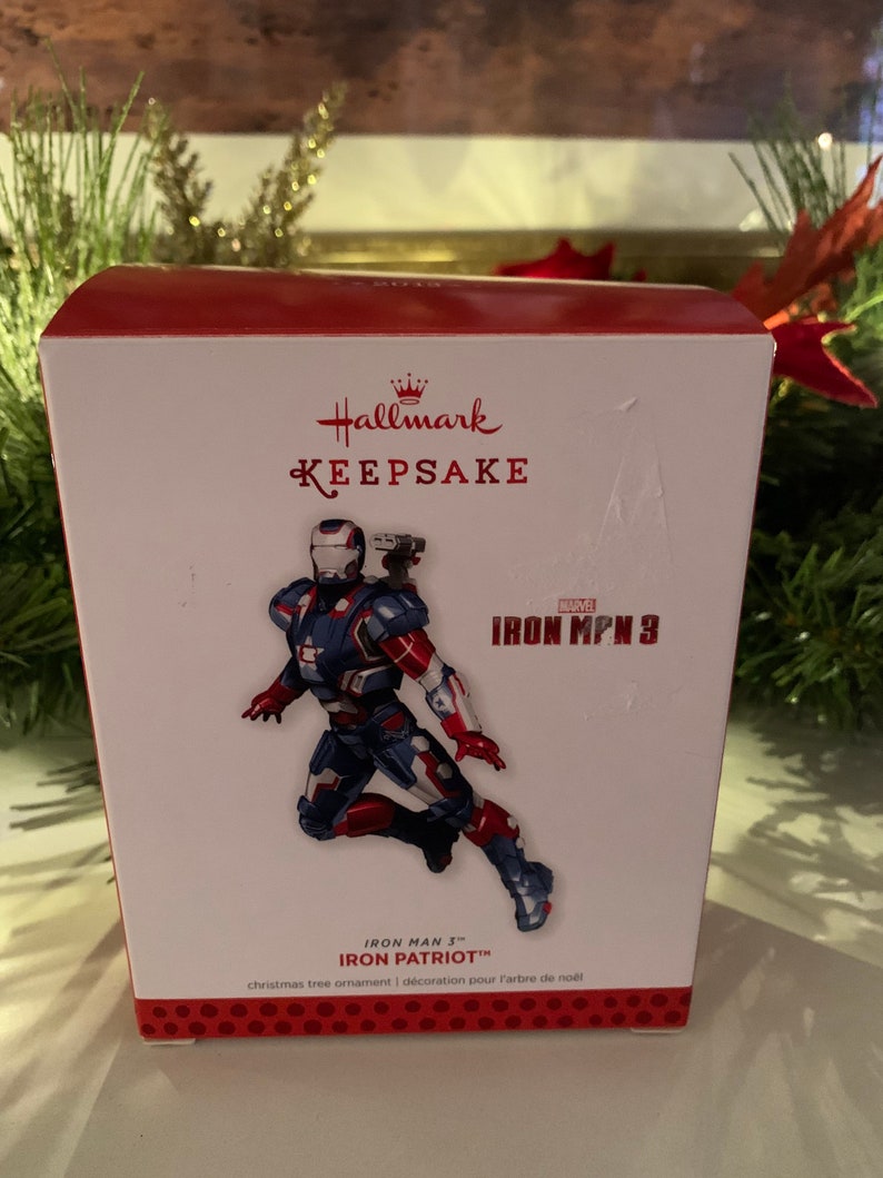 May include: Red and white Hallmark Keepsake ornament box featuring Iron Patriot from Iron Man 3. The ornament is a Christmas tree decoration.