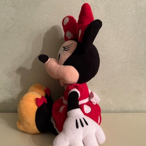 Op de afbeelding: Een pluche speelgoed van Minnie Mouse die zit met haar benen gekruist. Ze draagt een rode jurk met witte stippen en een rode strik met witte stippen. Haar oren zijn zwart en haar gezicht is roze met zwarte ogen en een zwarte neus.