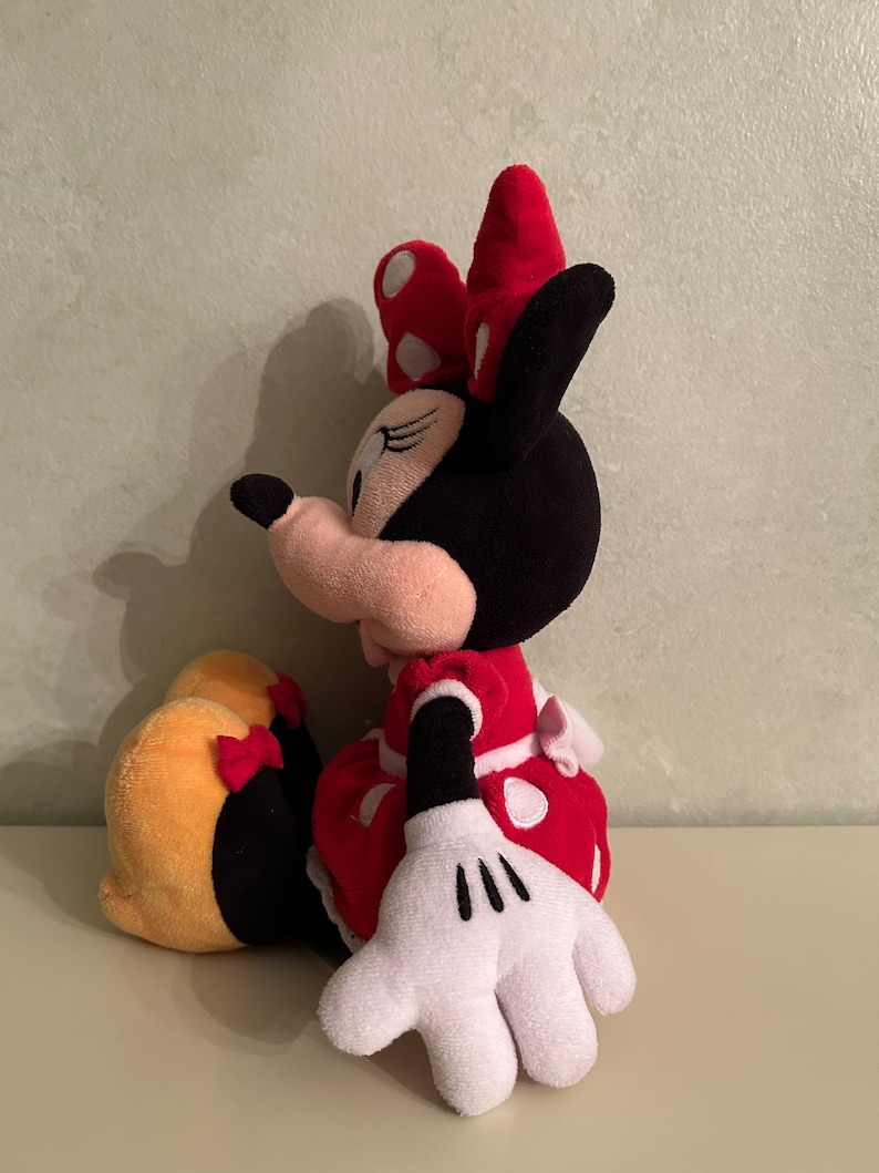 Op de afbeelding: Een pluche speelgoed van Minnie Mouse die zit met haar benen gekruist. Ze draagt een rode jurk met witte stippen en een rode strik met witte stippen. Haar oren zijn zwart en haar gezicht is roze met zwarte ogen en een zwarte neus.