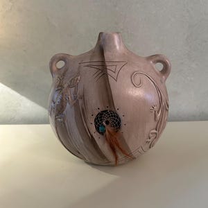 May include: A round, light brown ceramic vessel with two side handles, featuring embossed lizard figures, etched geometric lines, and a central circular cutout containing a small dreamcatcher with a turquoise bead and brown feathers.