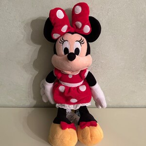 Op de afbeelding: Een rood en zwart pluche speelgoed van Minnie Mouse, die een rode jurk met witte stippen en een rode strik met witte stippen draagt.
