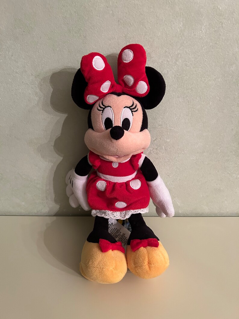 Op de afbeelding: Een rood en zwart pluche speelgoed van Minnie Mouse, die een rode jurk met witte stippen en een rode strik met witte stippen draagt.