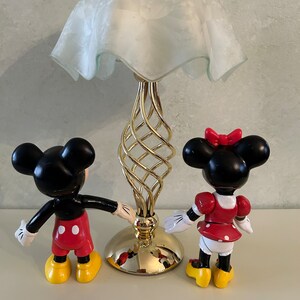 May include: A gold table lamp with a white frosted glass shade and a twisted base.  Two small plastic figurines of Mickey Mouse and Minnie Mouse stand on either side of the lamp.