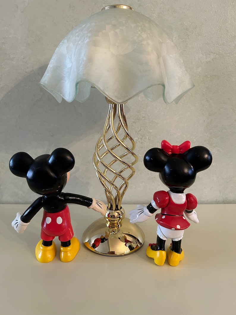 May include: A gold table lamp with a white frosted glass shade and a twisted base.  Two small plastic figurines of Mickey Mouse and Minnie Mouse stand on either side of the lamp.