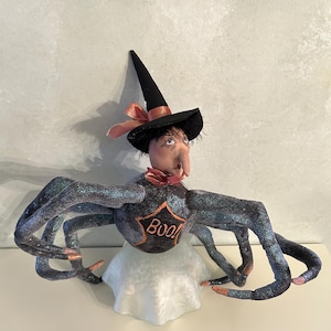May include: A black witch with a pointed hat and a star-shaped body with the word "Boo!" on it. The witch has eight long, glittery legs and is sitting on a white base.