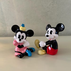 May include: Two ceramic figurines of cartoon characters. One is a female character in a pink dress with white polka dots, blue shoes, and a yellow hat. The other is a male character in red shorts, yellow shoes, and white gloves.