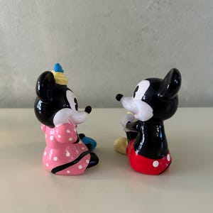 May include: Two ceramic figurines of Mickey and Minnie Mouse. Minnie is wearing a pink dress with white polka dots and a blue hat. Mickey is wearing red shorts with white polka dots and yellow shoes. Both figurines are black, white, and red.