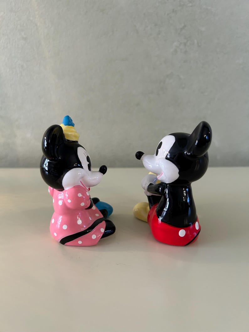 May include: Two ceramic figurines of Mickey and Minnie Mouse. Minnie is wearing a pink dress with white polka dots and a blue hat. Mickey is wearing red shorts with white polka dots and yellow shoes. Both figurines are black, white, and red.