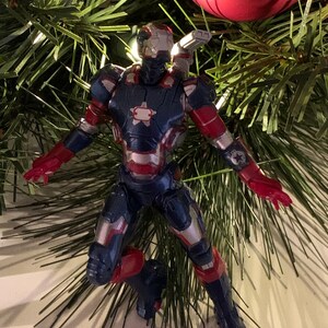 May include: A red, white and blue Iron Man action figure ornament hanging on a Christmas tree. The figure is in a flying pose with its arms outstretched.