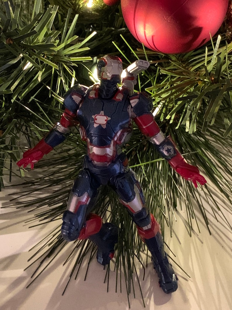 May include: A red, white and blue Iron Man action figure ornament hanging on a Christmas tree. The figure is in a flying pose with its arms outstretched.