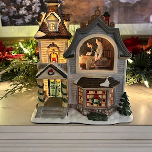 May include: A lighted Christmas village house with a detailed design. The house features a steeple, a front porch, and a bay window. The house has a brown roof with snow accents and a light inside. The house has a Christmas wreath on the front.