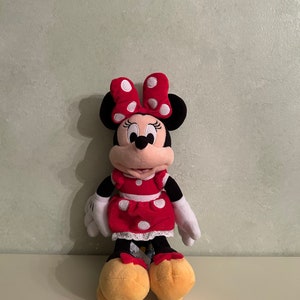 Op de afbeelding: Een rood en wit gestippeld pluche speelgoed van Minnie Mouse. Ze draagt een rode jurk met witte stippen en een witte strik met rode stippen. Ze heeft zwarte ogen en een zwarte neus.