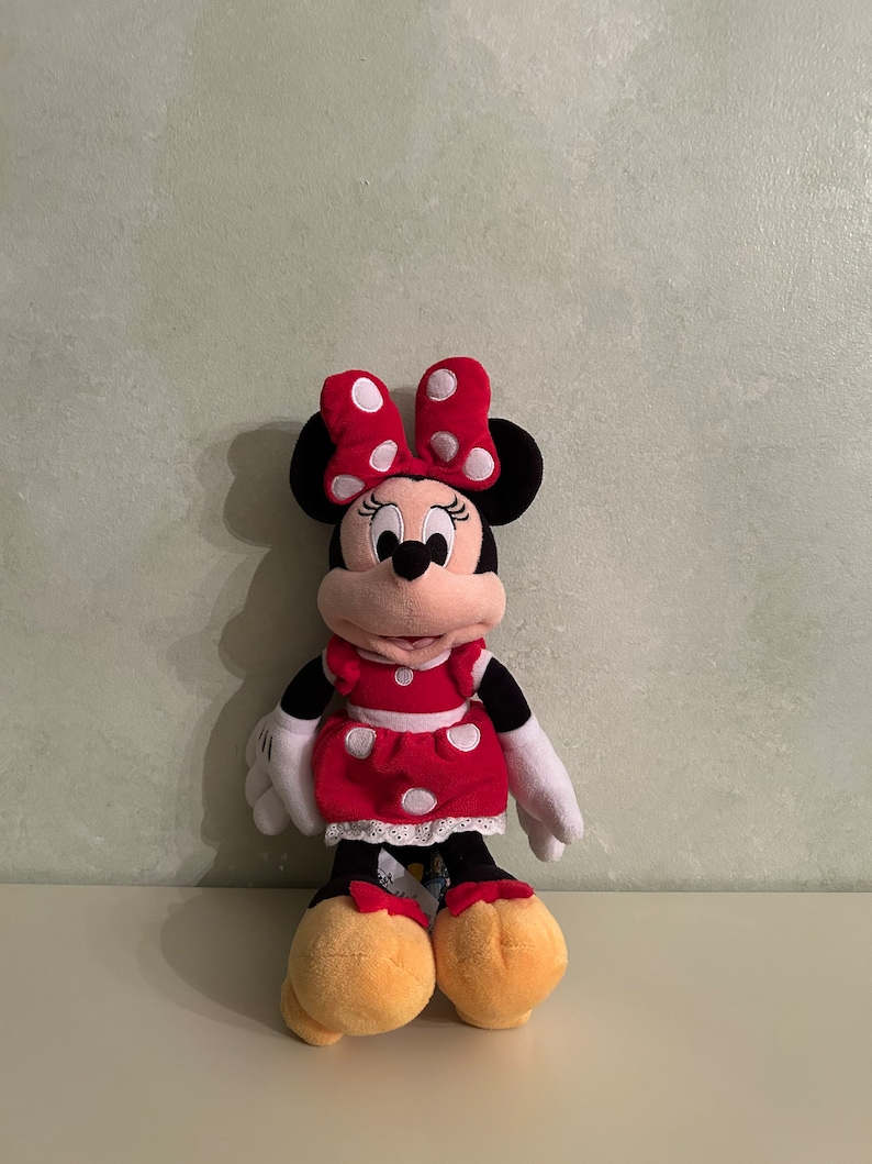 Op de afbeelding: Een rood en wit gestippeld pluche speelgoed van Minnie Mouse. Ze draagt een rode jurk met witte stippen en een witte strik met rode stippen. Ze heeft zwarte ogen en een zwarte neus.