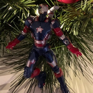 May include: A red, white and blue action figure of Iron Man in a flying pose. The figure is a Christmas ornament and is hanging from a Christmas tree.